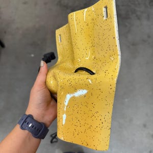 K&ouml;nnte beinhalten: Eine gl&auml;nzende gelbe Wandleuchte aus Keramik mit schwarzem Sprenkelmuster. Die Leuchte hat eine geschwungene, skulpturale Form mit zwei rechteckigen Befestigungsschlitzen an der Oberseite und einer zentralen runden &Ouml;ffnung f&uuml;r ein schwarzes Stromkabel.