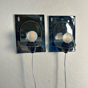 May include: Two wall-mounted sconces with a dark blue and black finish. Each sconce features a curved, semi-circular design with a frosted globe light bulb. The sconces are connected to a wall outlet via a silver wire.