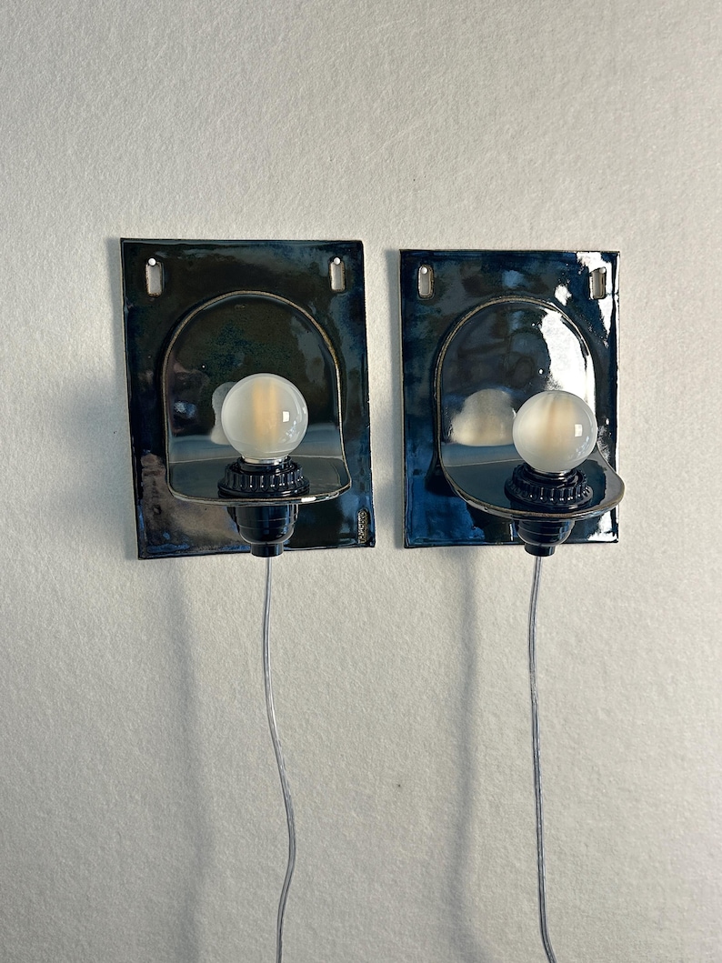 May include: Two wall-mounted sconces with a dark blue and black finish. Each sconce features a curved, semi-circular design with a frosted globe light bulb. The sconces are connected to a wall outlet via a silver wire.