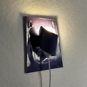 Pode incluir: Uma lumin&aacute;ria de parede moderna com uma placa traseira retangular roxa escura brilhante. A lumin&aacute;ria apresenta uma sombra curva roxa escura e uma l&acirc;mpada redonda e brilhante. Um cabo de alimenta&ccedil;&atilde;o transparente se estende da lumin&aacute;ria.