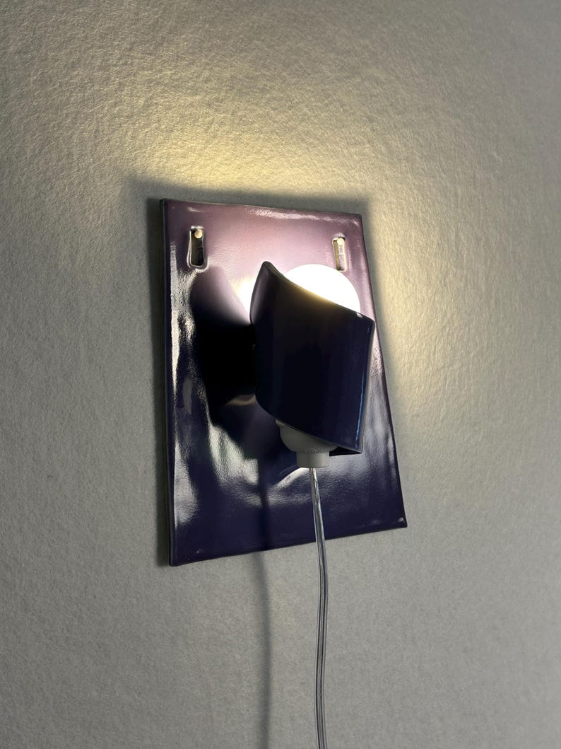 Pode incluir: Uma lumin&aacute;ria de parede moderna com uma placa traseira retangular roxa escura brilhante. A lumin&aacute;ria apresenta uma sombra curva roxa escura e uma l&acirc;mpada redonda e brilhante. Um cabo de alimenta&ccedil;&atilde;o transparente se estende da lumin&aacute;ria.