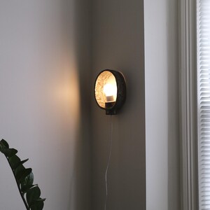 May include: A round, dark-colored wall sconce with a textured interior and a lit bulb. The sconce is mounted on a gray wall, with a cord hanging down. A plant is visible in the lower left corner.