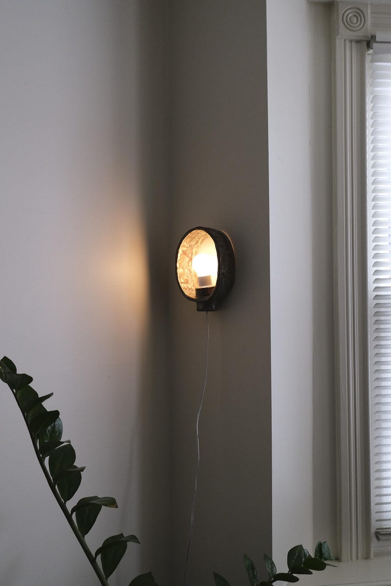 May include: A round, dark-colored wall sconce with a textured interior and a lit bulb. The sconce is mounted on a gray wall, with a cord hanging down. A plant is visible in the lower left corner.