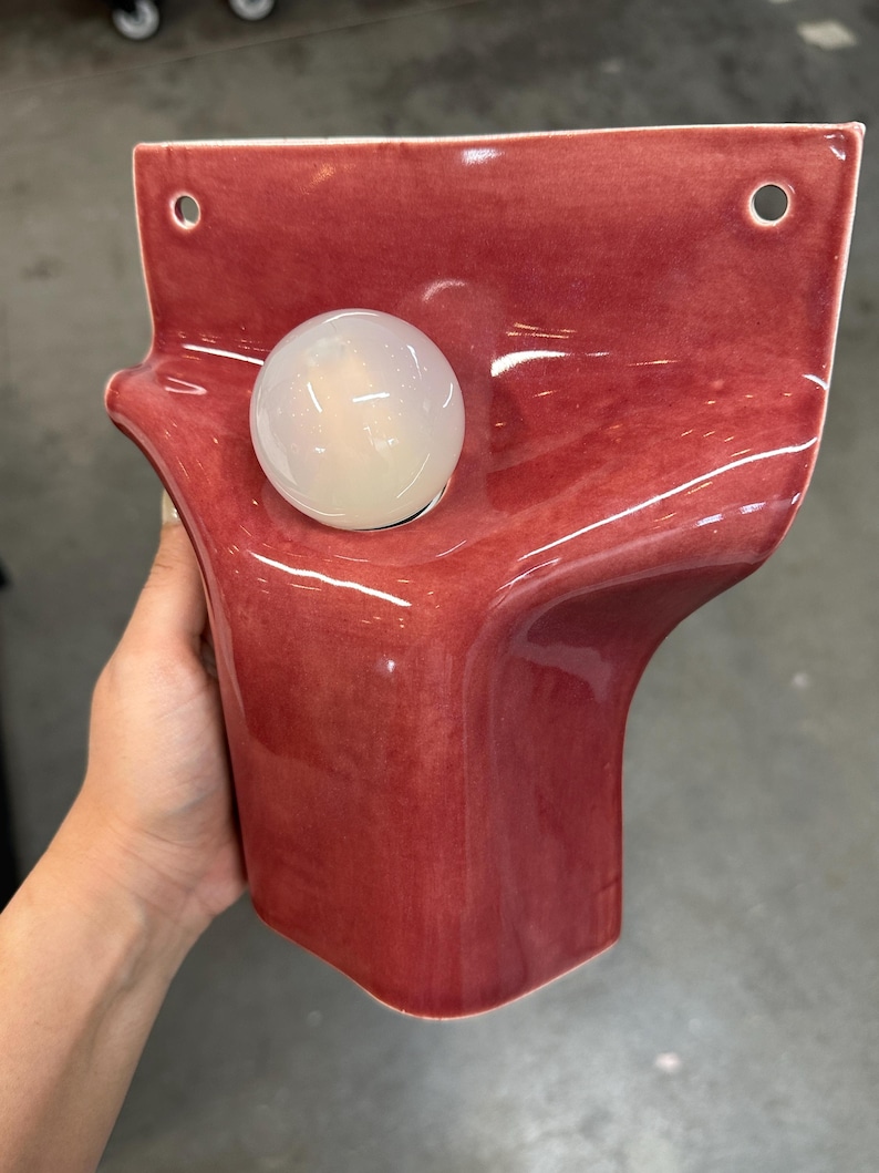 Pode incluir: Um candeeiro de parede em cer&acirc;mica com um acabamento brilhante em rosa profundo e uma forma org&acirc;nica ondulada. A pe&ccedil;a inclui dois orif&iacute;cios de montagem circulares no topo e uma l&acirc;mpada esf&eacute;rica &uacute;nica em vidro branco fosco.