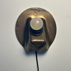 May include: A wall-mounted lamp featuring a circular, metallic bronze-toned backplate with a folded, hat-like shape. A black socket holds a single, round, frosted white light bulb. A thin black power cord extends downward from the base of the fixture.
