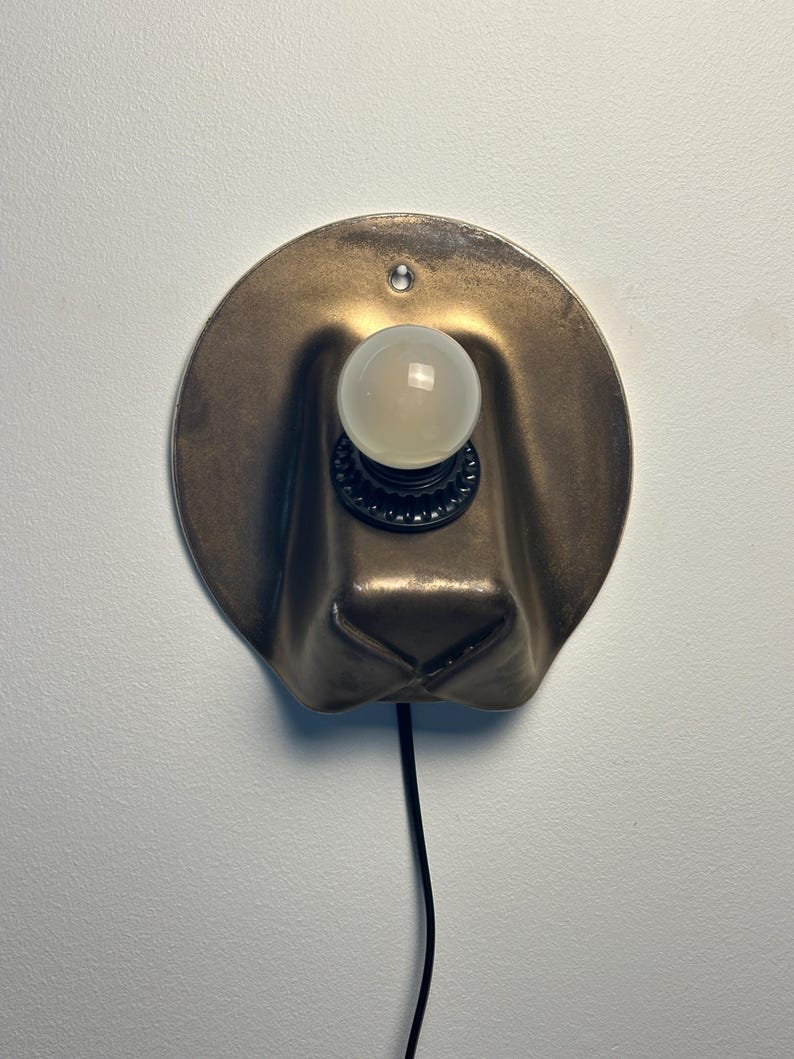 May include: A wall-mounted lamp featuring a circular, metallic bronze-toned backplate with a folded, hat-like shape. A black socket holds a single, round, frosted white light bulb. A thin black power cord extends downward from the base of the fixture.