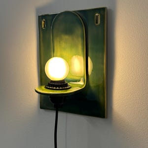 May include: A wall-mounted ceramic sconce featuring a glossy emerald green finish with a rectangular backplate and an arched center. A small, round, frosted white light bulb sits on a protruding shelf, connected by a black cord and base.