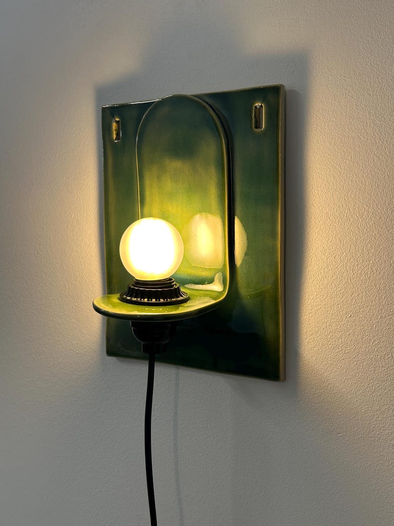 May include: A wall-mounted ceramic sconce featuring a glossy emerald green finish with a rectangular backplate and an arched center. A small, round, frosted white light bulb sits on a protruding shelf, connected by a black cord and base.