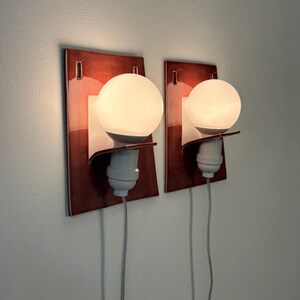 Puede incluir: Dos apliques de pared con placas traseras rectangulares de cer&aacute;mica roja oscura brillante. Cada aplique tiene un soporte blanco curvado que sujeta un casquillo blanco y una bombilla redonda de cristal esmerilado, con un cable gris fino.