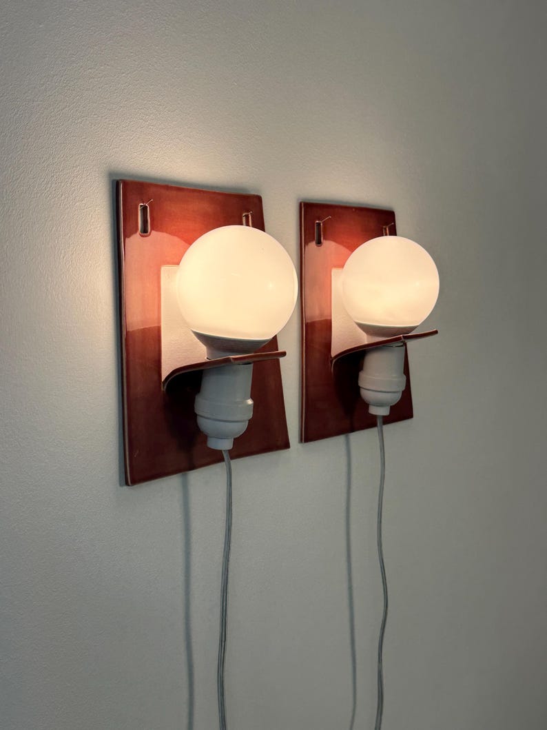 Puede incluir: Dos apliques de pared con placas traseras rectangulares de cer&aacute;mica roja oscura brillante. Cada aplique tiene un soporte blanco curvado que sujeta un casquillo blanco y una bombilla redonda de cristal esmerilado, con un cable gris fino.