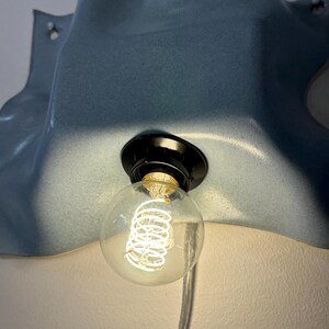 May include: A wall-mounted light fixture featuring a smooth, blue, wavy ceramic shade with a glossy finish. A black socket holds a clear, round globe light bulb with a visible glowing coiled filament. A transparent power cord hangs downward.