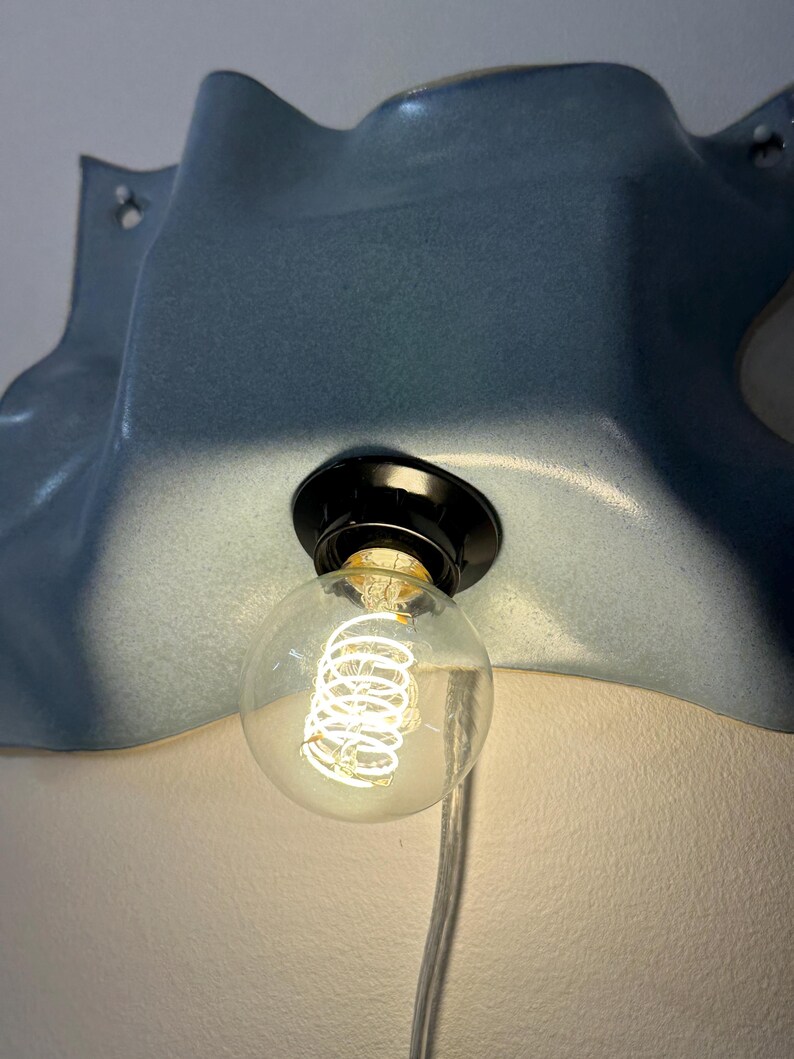 May include: A wall-mounted light fixture featuring a smooth, blue, wavy ceramic shade with a glossy finish. A black socket holds a clear, round globe light bulb with a visible glowing coiled filament. A transparent power cord hangs downward.