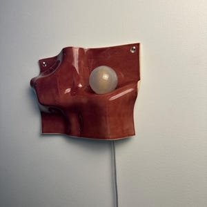 May include: A wall-mounted ceramic light fixture with a glossy, deep reddish-brown finish and an undulating, organic shape. The piece features a single spherical, frosted white light bulb nestled within a recessed curve and a thin power cord.