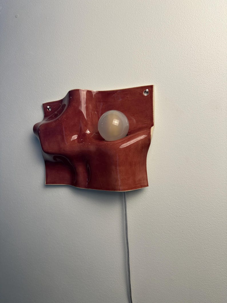 May include: A wall-mounted ceramic light fixture with a glossy, deep reddish-brown finish and an undulating, organic shape. The piece features a single spherical, frosted white light bulb nestled within a recessed curve and a thin power cord.