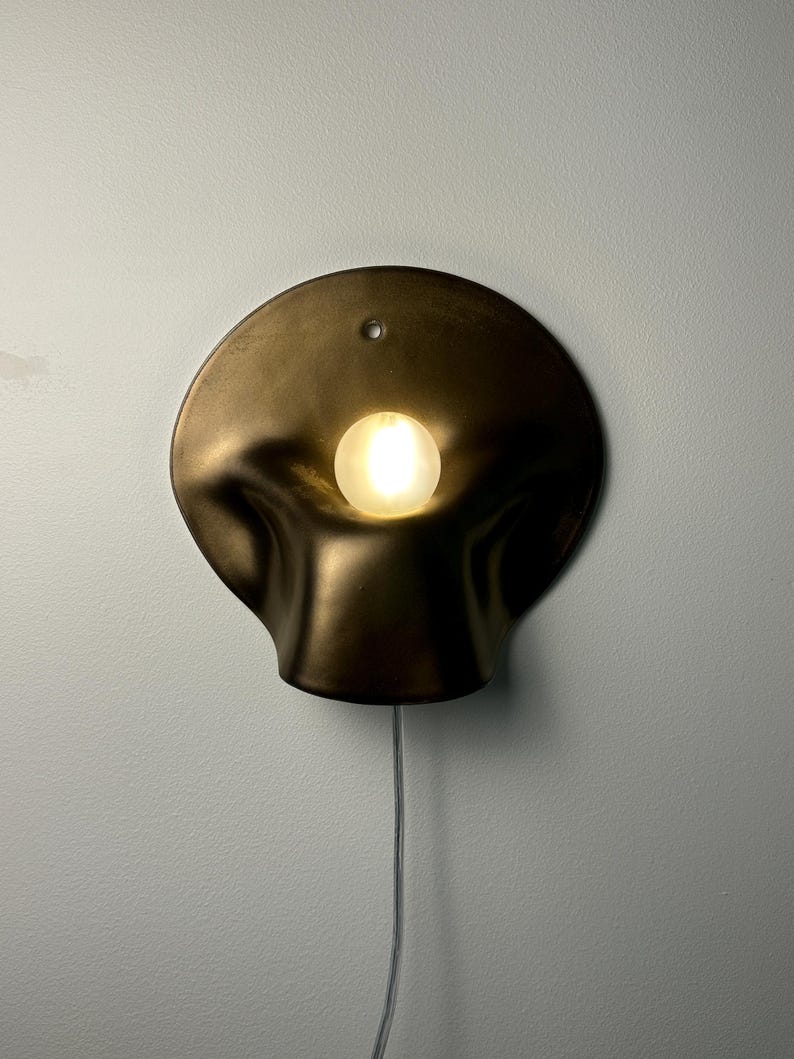 May include: A wall-mounted lamp featuring a dark, metallic, organic-shaped shade with a smooth, undulating surface. A single, glowing, spherical light bulb is positioned in the center of the shade. A thin, clear power cord hangs from the bottom.