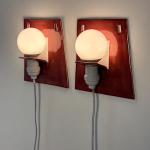 Puede incluir: Un par de apliques de pared con placas traseras de cer&aacute;mica de color rojo oscuro brillante y forma trapezoidal. Cada l&aacute;mpara incluye una bombilla esf&eacute;rica blanca, un casquillo blanco y un cable el&eacute;ctrico gris que baja desde la base.