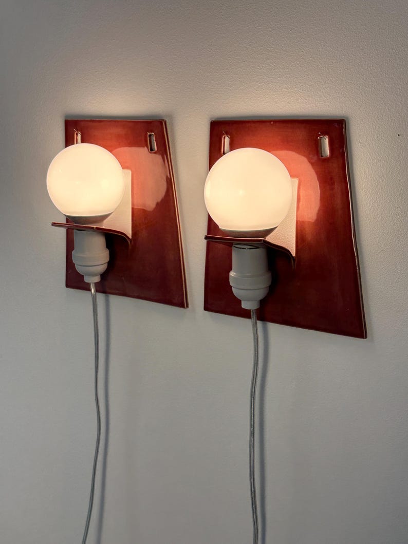 Puede incluir: Un par de apliques de pared con placas traseras de cer&aacute;mica de color rojo oscuro brillante y forma trapezoidal. Cada l&aacute;mpara incluye una bombilla esf&eacute;rica blanca, un casquillo blanco y un cable el&eacute;ctrico gris que baja desde la base.