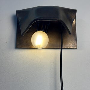 Pode incluir: Um candeeiro de parede com um abajur de metal escuro e curvo com acabamento desgastado. Uma l&acirc;mpada redonda e fosca est&aacute; centrada sob o abajur, emitindo uma luz quente. Um cabo de alimenta&ccedil;&atilde;o preto pende verticalmente do candeeiro.