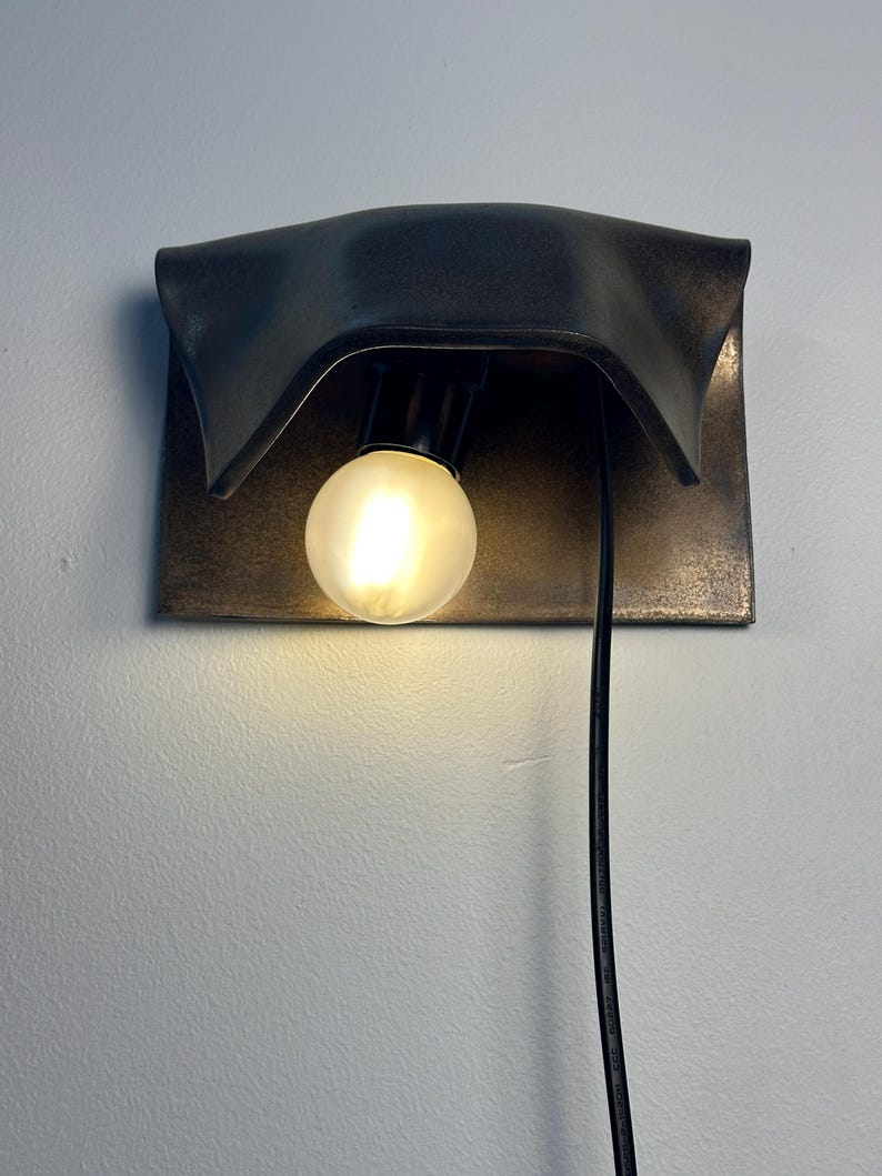 Pode incluir: Um candeeiro de parede com um abajur de metal escuro e curvo com acabamento desgastado. Uma l&acirc;mpada redonda e fosca est&aacute; centrada sob o abajur, emitindo uma luz quente. Um cabo de alimenta&ccedil;&atilde;o preto pende verticalmente do candeeiro.