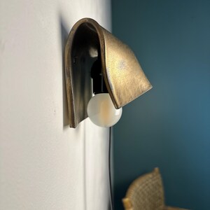 Pode incluir: Um candeeiro de parede com um abajur curvo em metal tom lat&atilde;o, com um acabamento texturizado e envelhecido. A lumin&aacute;ria inclui um casquilho preto com uma l&acirc;mpada redonda de vidro fosco branco e um fio preto fino que desce.