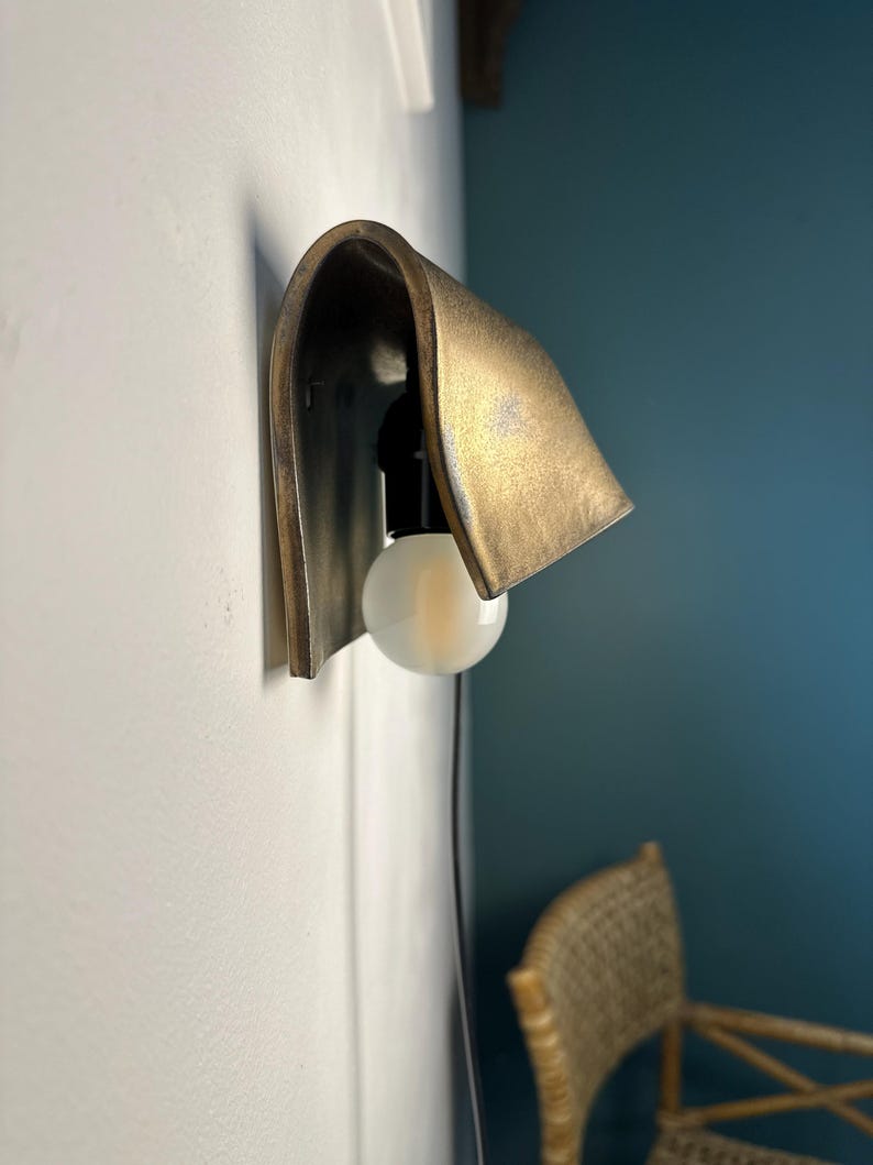 Pode incluir: Um candeeiro de parede com um abajur curvo em metal tom lat&atilde;o, com um acabamento texturizado e envelhecido. A lumin&aacute;ria inclui um casquilho preto com uma l&acirc;mpada redonda de vidro fosco branco e um fio preto fino que desce.