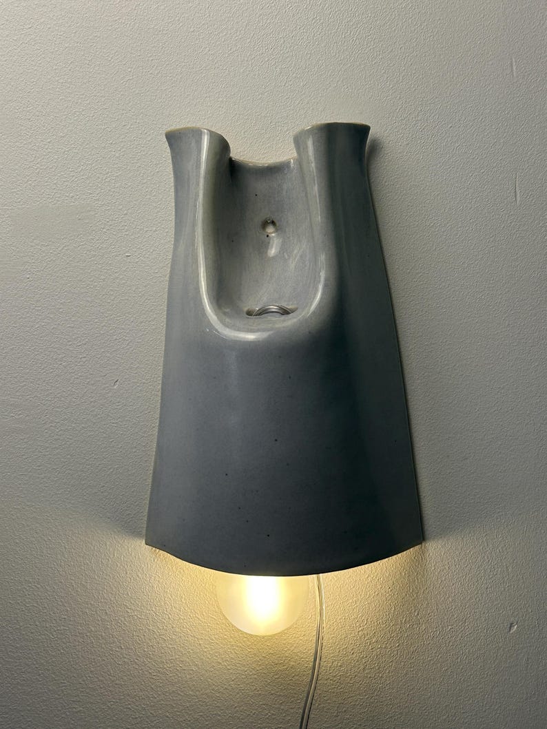 K&ouml;nnte beinhalten: Eine Wandleuchte aus Keramik mit einer glatten, gl&auml;nzenden hellgrauen Oberfl&auml;che. Die Leuchte hat eine hohe, geschwungene U-Form mit einer zentralen &Ouml;ffnung f&uuml;r ein Leuchtmittel. Eine einzelne leuchtende Gl&uuml;hbirne ist unten sichtbar.