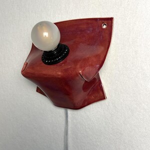 Puede incluir: Un aplique de pared con una base de cer&aacute;mica de color rojo intenso brillante con una forma org&aacute;nica y plegada. Un casquillo negro sostiene una bombilla redonda de color blanco esmerilado. Un cable transparente sale por abajo y se ve un agujero.