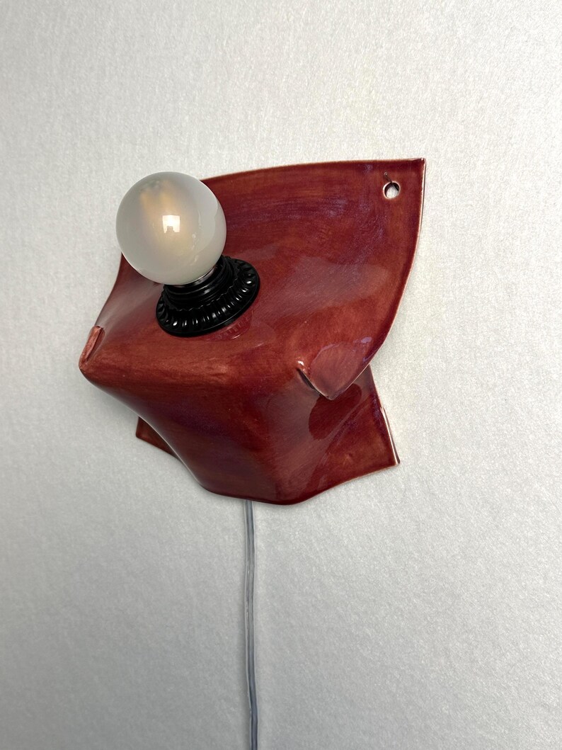 Puede incluir: Un aplique de pared con una base de cer&aacute;mica de color rojo intenso brillante con una forma org&aacute;nica y plegada. Un casquillo negro sostiene una bombilla redonda de color blanco esmerilado. Un cable transparente sale por abajo y se ve un agujero.