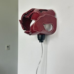 May include: A burgundy ceramic wall sconce with an abstract design. The sconce has a glossy finish and is mounted on a white wall with a black light fixture and a silver cord.