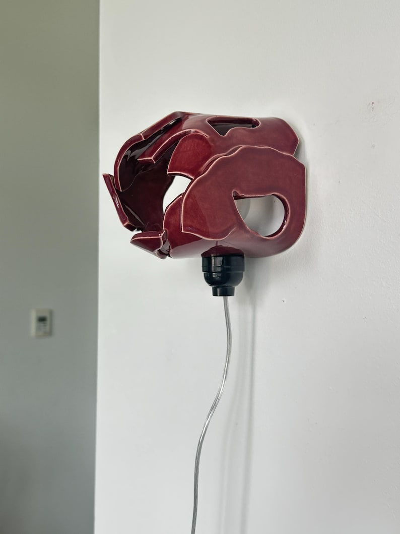 May include: A burgundy ceramic wall sconce with an abstract design. The sconce has a glossy finish and is mounted on a white wall with a black light fixture and a silver cord.