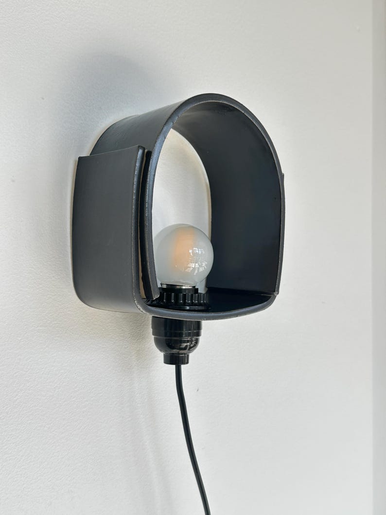 Puede incluir: Una l&aacute;mpara de pared negra con una pantalla curva en forma de arco. La l&aacute;mpara alberga una bombilla y est&aacute; conectada a un cable el&eacute;ctrico negro. La luz est&aacute; montada en una pared blanca.
