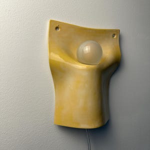 May include: A wall-mounted lamp featuring a curved, yellow ceramic shade with a smooth, glossy finish. The shade has two small circular holes at the top corners for mounting and holds a single, round, frosted white glass light bulb in the center.