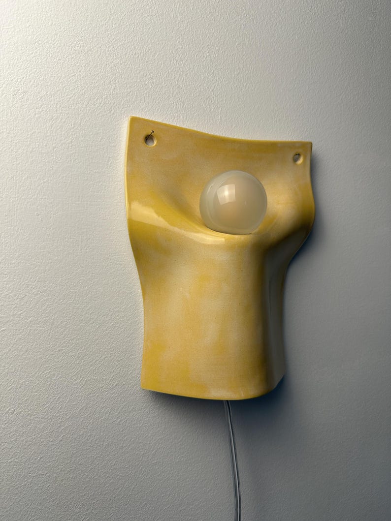 May include: A wall-mounted lamp featuring a curved, yellow ceramic shade with a smooth, glossy finish. The shade has two small circular holes at the top corners for mounting and holds a single, round, frosted white glass light bulb in the center.