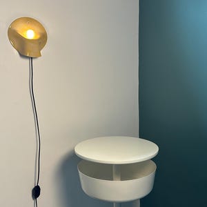 May include: A wall-mounted light fixture with a curved, pale yellow shade emitting a warm glow, connected by a black cord to a switch. Below it stands a white circular side table featuring a flat top surface and a deep, cylindrical storage compartment.
