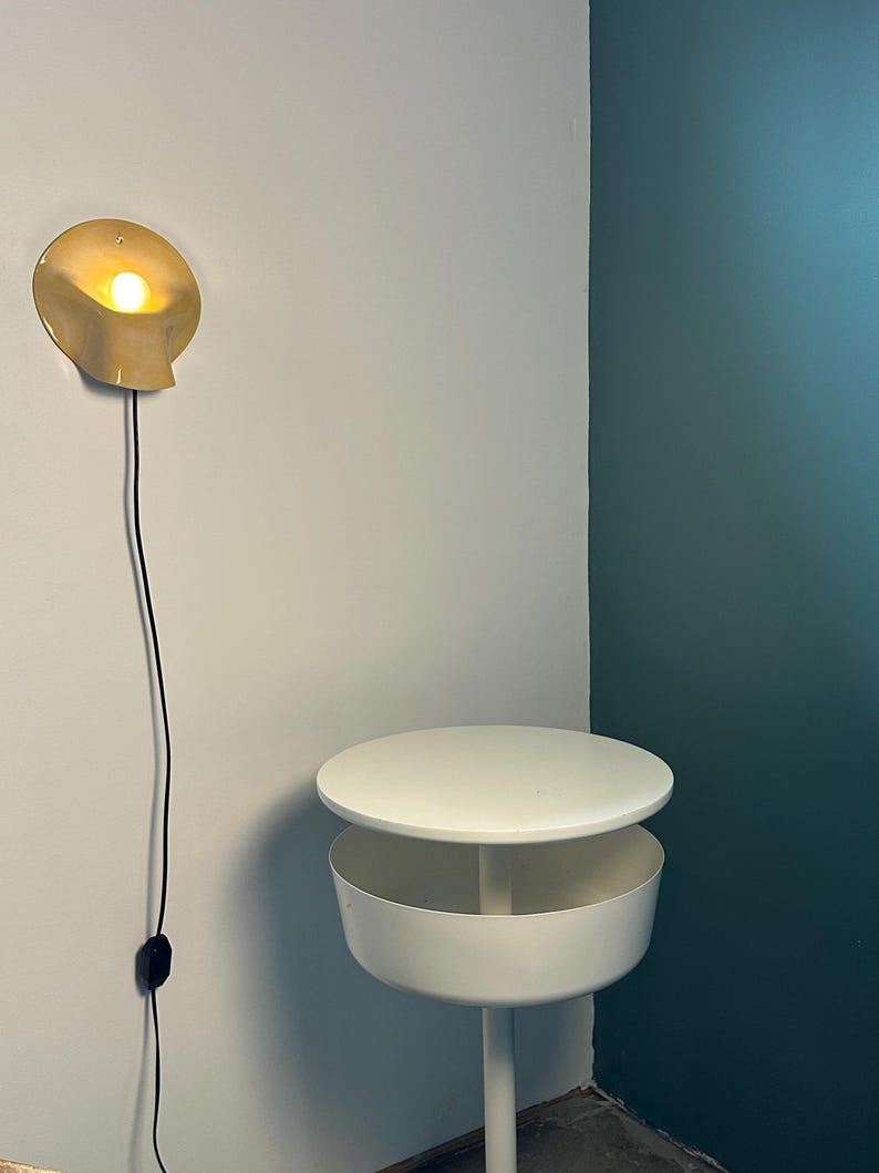 May include: A wall-mounted light fixture with a curved, pale yellow shade emitting a warm glow, connected by a black cord to a switch. Below it stands a white circular side table featuring a flat top surface and a deep, cylindrical storage compartment.