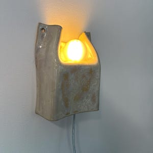 May include: A wall-mounted ceramic sconce with a glossy, off-white glaze and mottled beige patches. The fixture features a U-shaped cutout at the top, housing a single glowing, warm-toned light bulb. A thin, light-coloured power cord extends downwards.