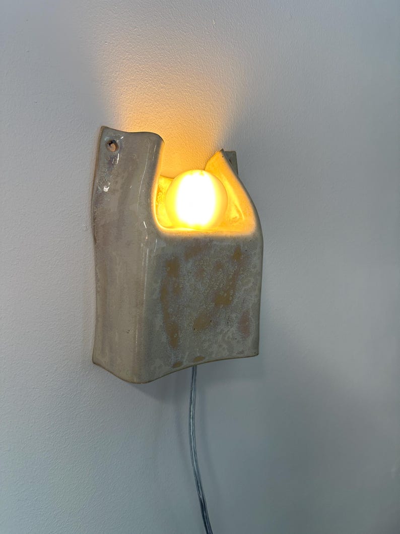 May include: A wall-mounted ceramic sconce with a glossy, off-white glaze and mottled beige patches. The fixture features a U-shaped cutout at the top, housing a single glowing, warm-toned light bulb. A thin, light-coloured power cord extends downwards.