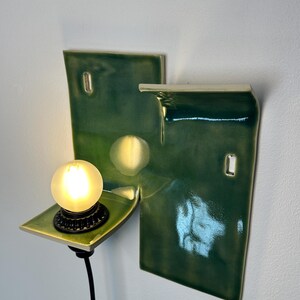 May include: A wall-mounted ceramic light fixture featuring a glossy, deep green glazed finish. The structure consists of two rectangular panels with a small, protruding shelf holding a round, glowing light bulb and a black cord extending downward.