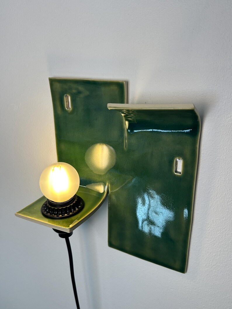 May include: A wall-mounted ceramic light fixture featuring a glossy, deep green glazed finish. The structure consists of two rectangular panels with a small, protruding shelf holding a round, glowing light bulb and a black cord extending downward.