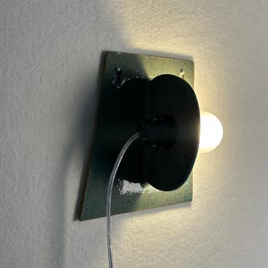 May include: A wall-mounted lamp featuring a dark teal, speckled ceramic square backplate with a circular center piece. A single glowing light bulb extends from the side, connected by a clear electrical cord that hangs down from the fixture.