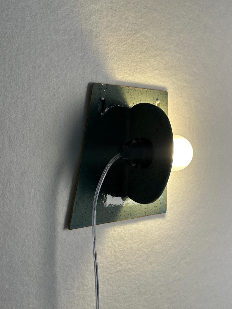 May include: A wall-mounted lamp featuring a dark teal, speckled ceramic square backplate with a circular center piece. A single glowing light bulb extends from the side, connected by a clear electrical cord that hangs down from the fixture.