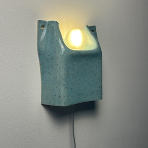 May include: A wall-mounted light fixture featuring a light blue ceramic housing with a dark speckled pattern. The structure has a U-shaped cutout at the top that holds a single, glowing, spherical white light bulb. A thin power cord hangs below.