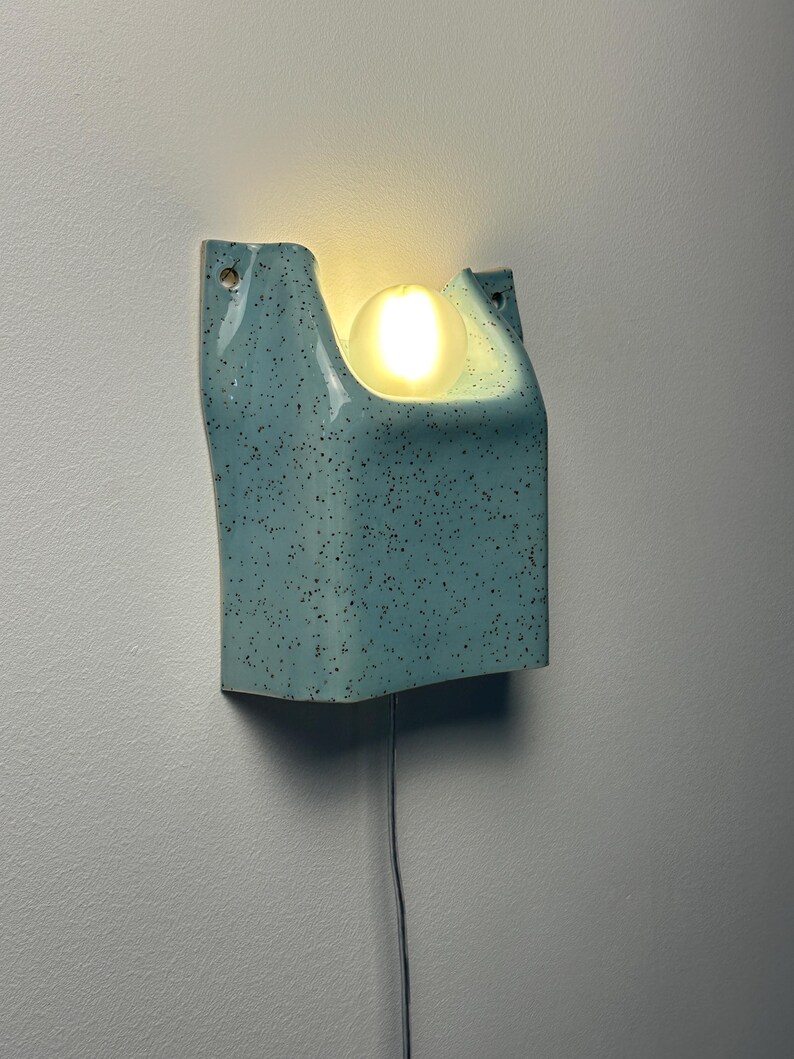 May include: A wall-mounted light fixture featuring a light blue ceramic housing with a dark speckled pattern. The structure has a U-shaped cutout at the top that holds a single, glowing, spherical white light bulb. A thin power cord hangs below.