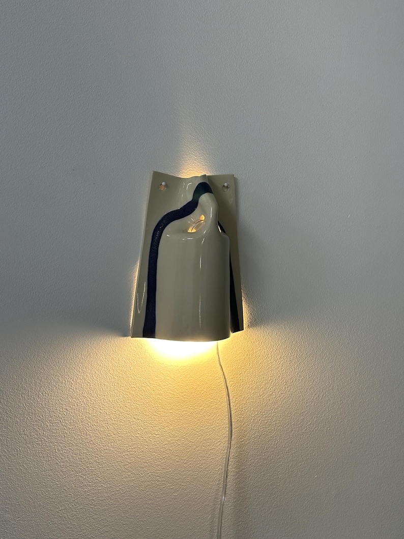 May include: A wall-mounted ceramic light fixture with a glossy off-white finish and a dark blue vertical stripe. The sconce features a curved, folded design that emits a warm, glowing light from both the top and bottom openings, with a clear cord.