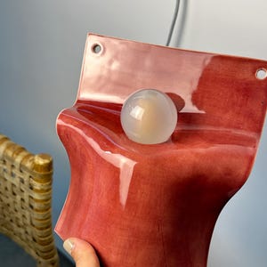 May include: A wall-mounted light fixture featuring a glossy, deep red ceramic base with a curved, wave-like shape. The fixture includes a circular, translucent white glass globe bulb centered on the ceramic surface and two mounting holes at the top.