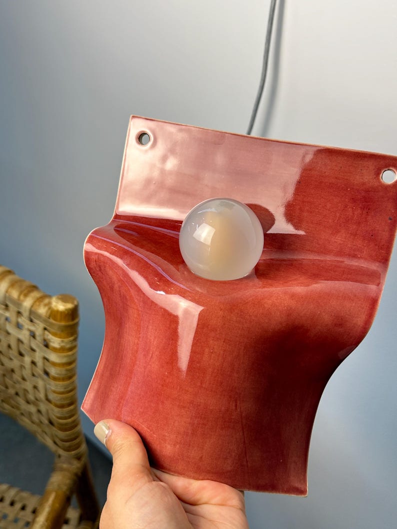 May include: A wall-mounted light fixture featuring a glossy, deep red ceramic base with a curved, wave-like shape. The fixture includes a circular, translucent white glass globe bulb centered on the ceramic surface and two mounting holes at the top.