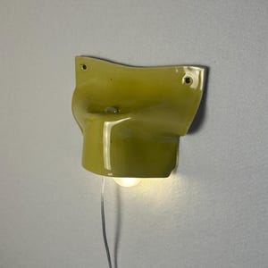 May include: A chartreuse-colored wall sconce with a curved design. The sconce has a small light bulb and a silver wire. The fixture is mounted on a light gray wall.