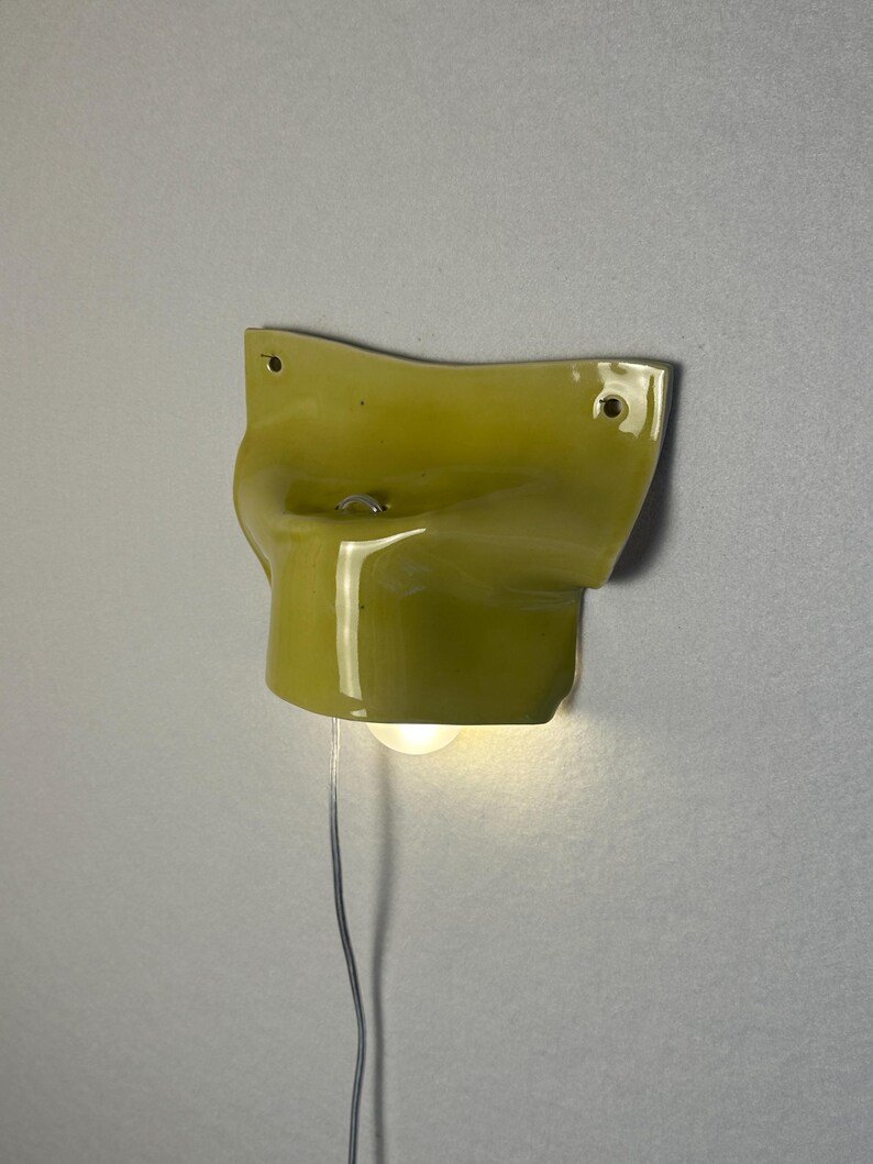 May include: A chartreuse-colored wall sconce with a curved design. The sconce has a small light bulb and a silver wire. The fixture is mounted on a light gray wall.