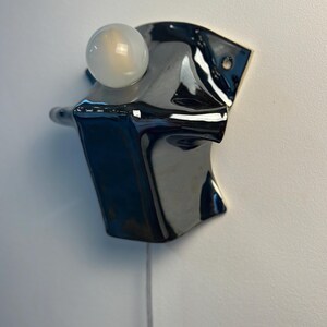 May include: A wall-mounted lamp featuring a glossy black ceramic base with an irregular, folded shape. A single, round, frosted white light bulb is positioned at the top of the fixture. A thin, light-colored electrical cord hangs from the bottom.