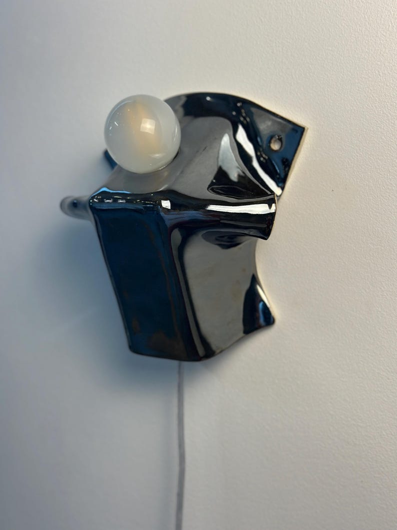 May include: A wall-mounted lamp featuring a glossy black ceramic base with an irregular, folded shape. A single, round, frosted white light bulb is positioned at the top of the fixture. A thin, light-colored electrical cord hangs from the bottom.